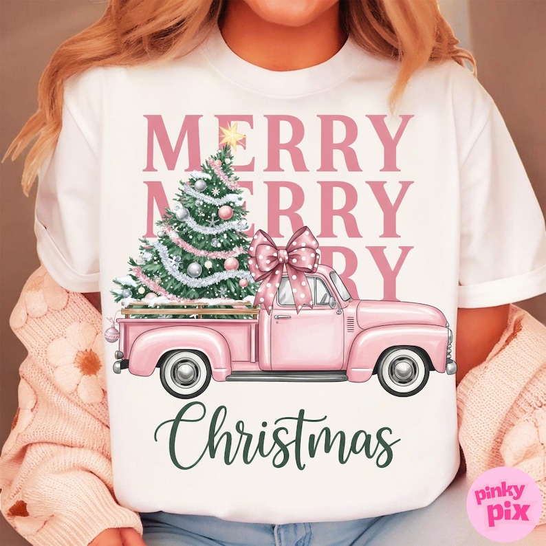May include: White t-shirt featuring a pink pickup truck carrying a Christmas tree and a large bow. The words "MERRY CHRISTMAS" are printed in pink and green. The design is festive and seasonal.
