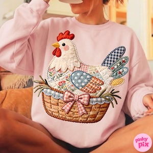 May include: A light pink sweatshirt featuring a patchwork chicken design. The chicken is in a basket with a gingham bow. The chicken has a red comb, yellow beak, and various patterned patches in blue, orange, and floral prints.