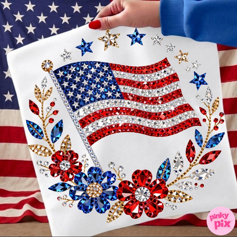 May include: A white square with a sparkling American flag design, featuring red, white, and blue crystals. The flag is surrounded by floral and star embellishments. The image is held by a person with red nail polish.