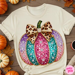 Op de afbeelding: Een cr&egrave;mekleurig t-shirt met een grote afbeelding van een pompoen versierd met veelkleurige strass-steentjes in roze, turquoise en paarse tinten, geaccentueerd met zilveren stervormige studs en een strik met luipaardprint met strass-rand.