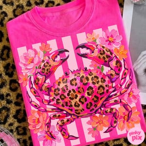 May include: A bright pink t-shirt featuring a crab design with leopard print accents. The crab is surrounded by pink and orange flowers and set against a background of white stripes. The shirt has the text "pinky pix" in the bottom right corner.