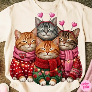 May include: A cream-colored t-shirt featuring a graphic of four smiling cats. The cats wear red and green sweaters, pink scarves, and heart-shaped headbands. One scarf has the word "LOVE".