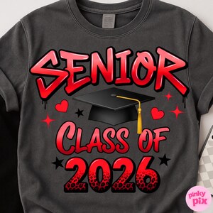 May include: Dark gray sweatshirt with the words "SENIOR CLASS OF 2026" in red graffiti-style lettering. A black graduation cap with a gold tassel is centered above the text, with red hearts and stars scattered around the design.