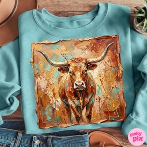 May include: A light blue sweatshirt featuring a detailed, textured painting of a longhorn. The artwork is predominantly brown, with accents of orange, yellow, and turquoise. The longhorn's horns are prominently displayed, and the overall style is rustic.