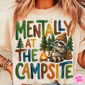 May include: Cream-colored t-shirt with the text "MENTALLY AT THE CAMPSITE" in colorful letters. A raccoon wearing a knit cap and holding a mug sits near a campfire and pine trees. A backpack is next to the raccoon.