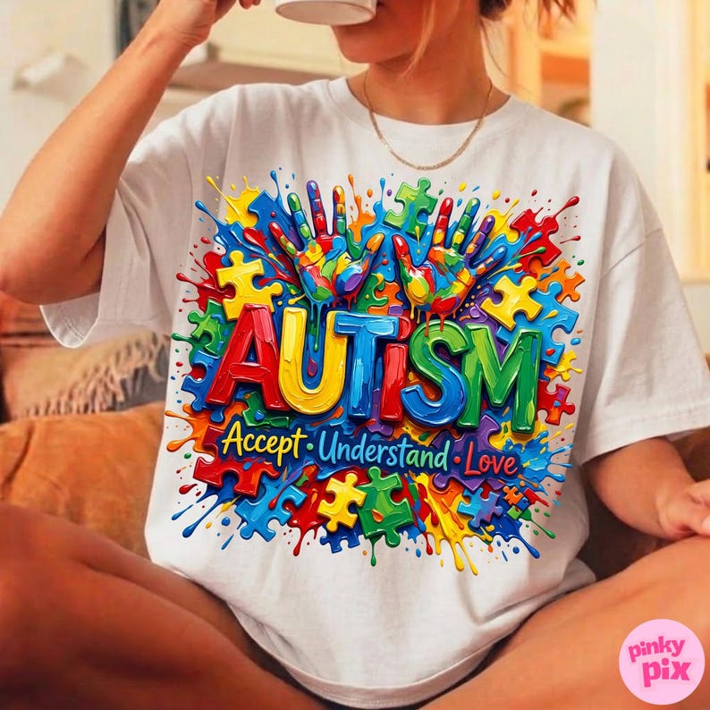 Op de afbeelding: Wit T-shirt met een kleurrijk grafisch ontwerp. Het ontwerp toont het woord "AUTISME" in kleurrijke letters, omringd door puzzelstukjes en handafdrukken. De woorden "Accepteer, Begrijp, Liefde" staan eronder.