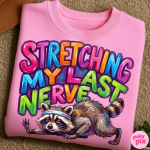 May include: Pink sweatshirt with the text "STRETCHING MY LAST NERVE" in colorful bubble letters. A raccoon wearing a headband is stretching below the text. The sweatshirt is a crew neck style.