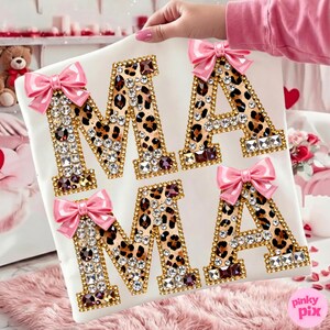 May include: White square cushion featuring the word "MAMA" in leopard print letters. Each letter is outlined in gold and decorated with clear and burgundy rhinestones. Pink bows are attached to the top of each letter.