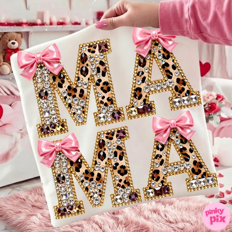 May include: White square cushion featuring the word "MAMA" in leopard print letters. Each letter is outlined in gold and decorated with clear and burgundy rhinestones. Pink bows are attached to the top of each letter.