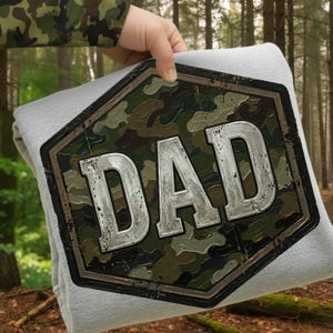 Pode incluir: Um cobertor cinza claro com um grande emblema com padr&atilde;o de camuflagem que diz "DAD" em letras prateadas. O emblema &eacute; emoldurado por uma borda marrom escura. O cobertor &eacute; segurado contra um fundo de floresta.