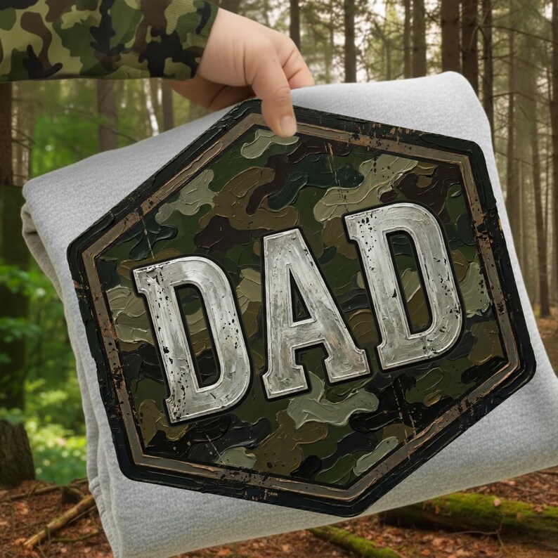 Pode incluir: Um cobertor cinza claro com um grande emblema com padr&atilde;o de camuflagem que diz "DAD" em letras prateadas. O emblema &eacute; emoldurado por uma borda marrom escura. O cobertor &eacute; segurado contra um fundo de floresta.