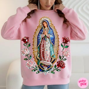 May include: A light pink sweatshirt featuring an image of the Virgin Mary with a jeweled halo and robe. The design includes red and pink roses with green stems and leaves. The sweatshirt has long sleeves and a crew neck.