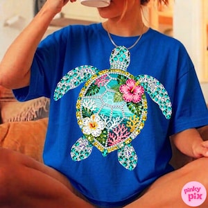 May include: Royal blue t-shirt featuring a detailed, bejeweled sea turtle design. The turtle's shell is adorned with turquoise, yellow, and clear gems, and it has a floral and coral design in the center. The shirt is a comfortable, casual style.
