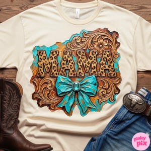 May include: Cream-coloured t-shirt with a decorative design featuring the word "MAMA" in leopard print. The design includes a turquoise bow and ornate, brown, and gold accents. A brown cowboy boot and denim jeans are visible.