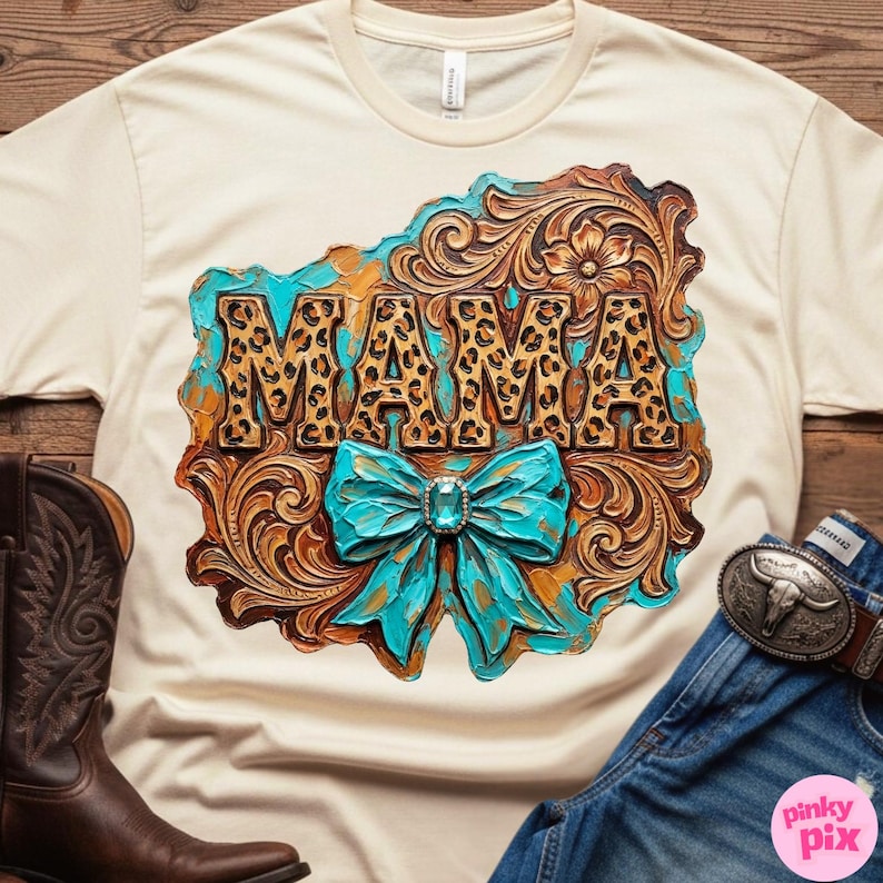 May include: Cream-colored t-shirt featuring a decorative design with the word "MAMA" in leopard print. The design includes a turquoise bow and ornate, brown, and gold accents. A brown cowboy boot and denim jeans are visible.