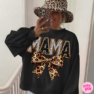 May include: Black sweatshirt with the word "MAMA" in jeweled letters and a leopard print bow. The sweatshirt has long sleeves. The person in the image is wearing a leopard print hat and holding a leopard print phone.