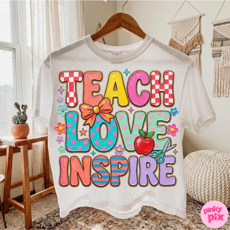 May include: White t-shirt with the words "TEACH LOVE INSPIRE" in colorful, bubble letters. The design includes an apple, scissors, and floral accents. The shirt is hanging on a wooden hanger.