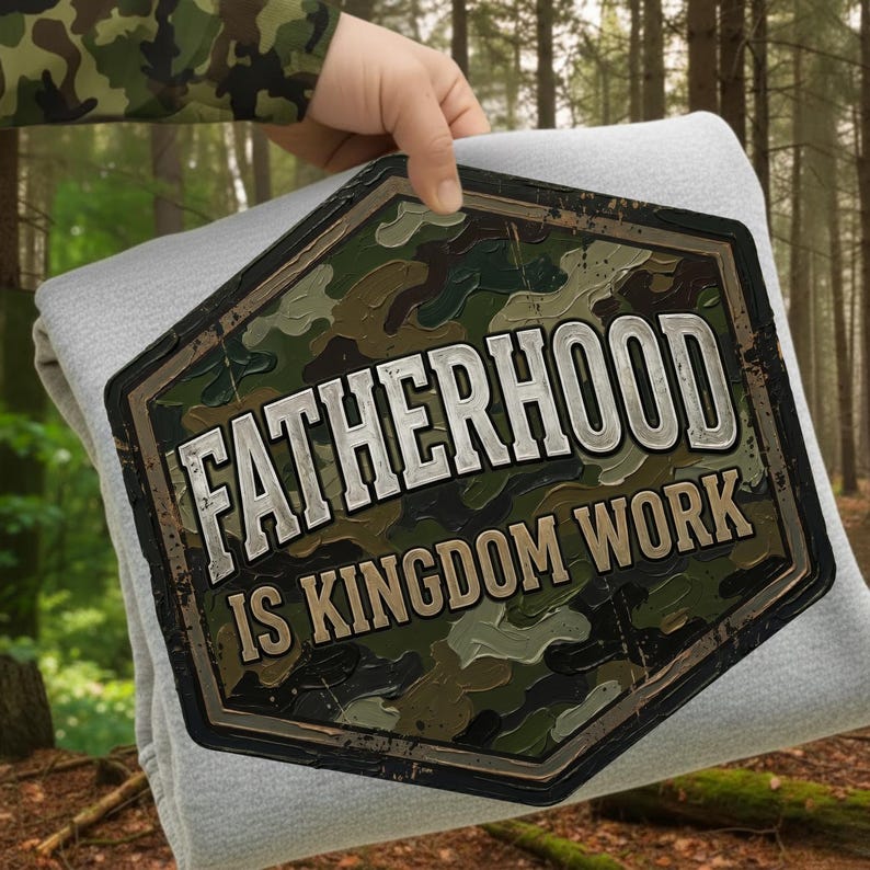 Pode incluir: Um cobertor cinza claro com um grande emblema com padr&atilde;o de camuflagem que diz "FATHERHOOD IS KINGDOM WORK" em letras brancas e marrons. O emblema tem uma forma hexagonal marrom escuro. O fundo &eacute; uma floresta.