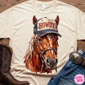 May include: Cream-colored t-shirt featuring a detailed illustration of a horse wearing a "HOWDY" cap. The horse has a brown coat, a bridle with turquoise accents, and a white mane. A pair of brown cowboy boots and blue jeans with a belt are visible.