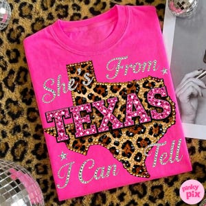 May include: A bright pink t-shirt featuring the text "She's From Texas I Can Tell". The design includes a leopard print Texas outline with the word "TEXAS" in sparkling pink letters. The shirt is a crew neck.