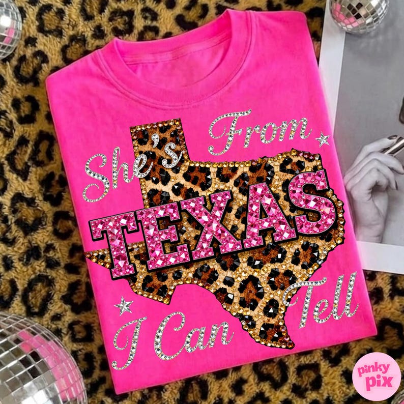 May include: A bright pink t-shirt featuring the text "She's From Texas I Can Tell". The design includes a leopard print Texas outline with the word "TEXAS" in sparkling pink letters. The shirt is a crew neck.