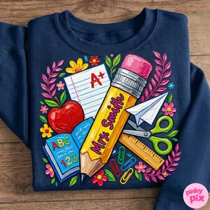 Pu&ograve; includere: Felpa blu navy con una grafica colorata a tema scolastico. Il design include una grande matita gialla con la scritta "Mrs. Smith", una mela, un libro aperto e altri materiali scolastici. Il design &egrave; circondato da fiori.