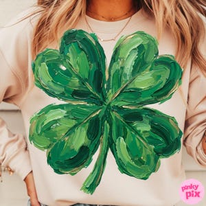 K&ouml;nnte beinhalten: Hellrosa Sweatshirt mit einem gro&szlig;en, strukturierten, gr&uuml;nen vierbl&auml;ttrigen Kleeblatt-Design. Das Kleeblatt hat goldene Akzente und ist mit sichtbaren Pinselstrichen bemalt. Das Sweatshirt ist ein l&auml;ssiger Stil, ideal f&uuml;r St. Patrick's Day.