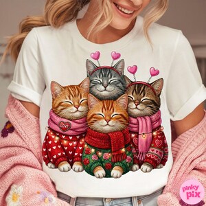 May include: White t-shirt featuring a graphic of four smiling cats. The cats wear sweaters, scarves, and heart-shaped headbands. The sweaters are red and green with heart and floral patterns. The scarves are pink and red.