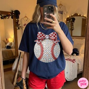 May include: Navy blue t-shirt featuring a baseball design with a sparkling, gem-studded baseball and a red and white bow. The baseball has red stitching. The person is wearing red shorts and a navy blue baseball cap. Baseball theme.
