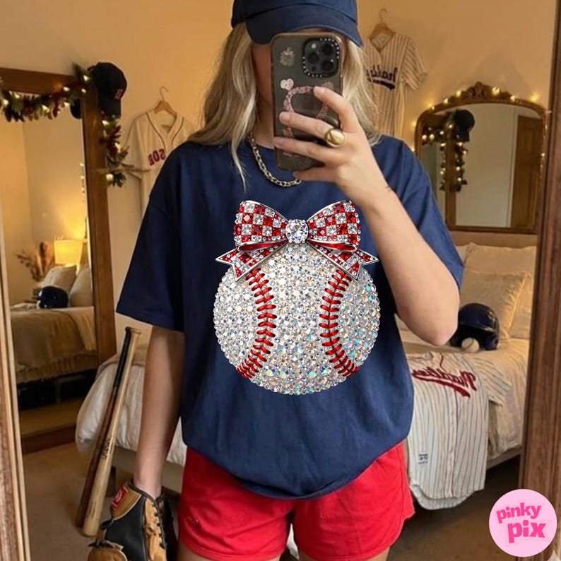 May include: Navy blue t-shirt featuring a baseball design with a sparkling, gem-studded baseball and a red and white bow. The baseball has red stitching. The person is wearing red shorts and a navy blue baseball cap. Baseball theme.