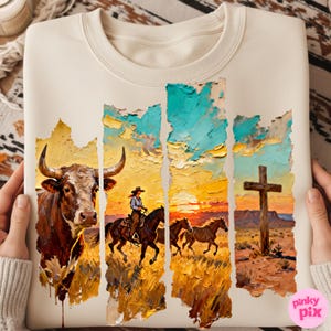 May include: Cream-colored sweatshirt featuring a Western-themed graphic. The design includes a bull, a cowgirl on horseback, running horses, and a cross against a sunset backdrop. The artwork is rendered in a textured, painterly style.