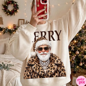 以下が含まれることがあります： クリーム色のクルーネックのスウェットシャツ。茶色の文字で「MERRY」と書かれ、その下にヒョウ柄のコート、お揃いのヒョウ柄の帽子、サングラス、大きなクリスタルのステートメントネックレスを身に着けたサンタクロースのグラフィックがプリントされています。