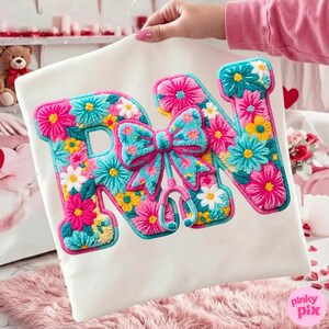 May include: White pillow with the letters "RN" embroidered with colorful flowers in pink, blue, yellow, and white. A bow and stethoscope are also embroidered. The pillow is held up by a person.
