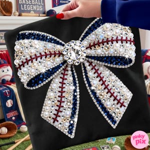 May include: Black pillow with a large, embellished bow design. The bow features white faux pearls, clear and blue crystals, and red stitching, resembling a baseball. A large, clear crystal is at the bow's center. The pillow is held by a person.