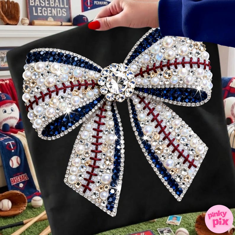 May include: Black pillow with a large, embellished bow design. The bow features white faux pearls, clear and blue crystals, and red stitching, resembling a baseball. A large, clear crystal is at the bow's center. The pillow is held by a person.