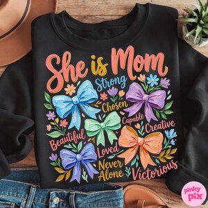 K&ouml;nnte beinhalten: Schwarzes Sweatshirt mit dem Text "She is Mom" in rosa und gelber Schrift. Das Design umfasst bunte Schleifen und florale Akzente mit W&ouml;rtern wie "Strong" und "Beautiful".