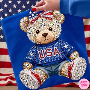 May include: A bedazzled teddy bear wearing a blue sweater with "USA" in red letters, denim shorts, and red, white, and blue sneakers. The bear has a bow with stars and stripes. The background includes a blue fabric and an American flag.