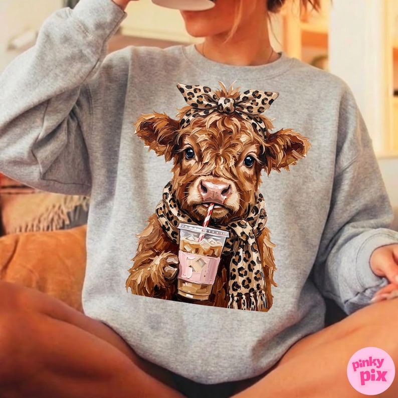 May include: A gray sweatshirt featuring a brown cow illustration. The cow wears a leopard print scarf and headband, and holds a coffee drink with a straw. The illustration has a cute and whimsical style.