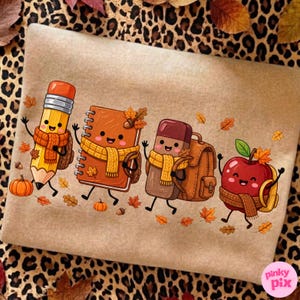 May include: A tan sweatshirt featuring a graphic of a cartoon pencil, notebook, eraser, and apple, each wearing a yellow scarf and carrying a backpack. The characters are surrounded by small orange pumpkins and scattered autumn leaves.