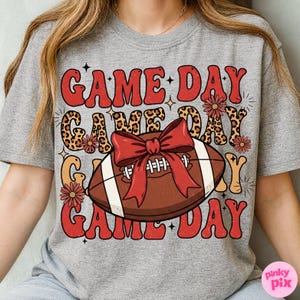 May include: Gray t-shirt with a retro design featuring the words "GAME DAY" in red and orange, with a football and bow graphic. The design includes leopard print accents and floral details, perfect for game day.