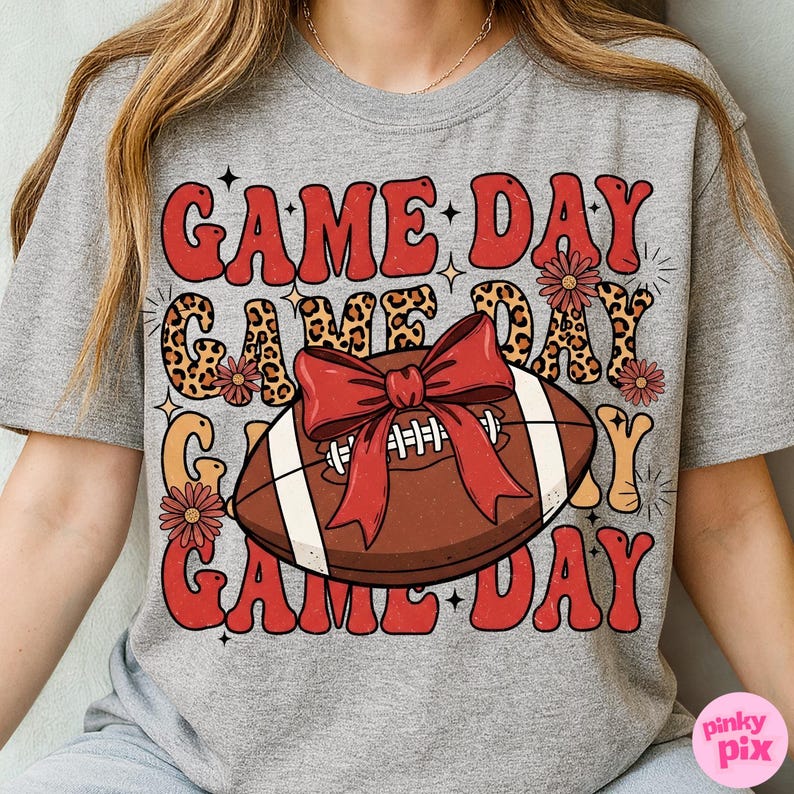 May include: Gray t-shirt with a retro design featuring the words "GAME DAY" in red and orange, with a football and bow graphic. The design includes leopard print accents and floral details, perfect for game day.