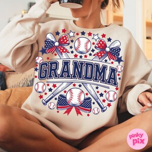 May include: A beige sweatshirt with a graphic design of two crossed baseball bats, baseballs, and red, white, and blue bows. The text "GRANDMA" is in the center of the design.
