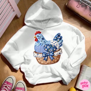 May include: White hooded sweatshirt featuring a colorful appliqu&eacute; of a blue chicken in a basket. The chicken design incorporates floral and polka dot patterns. The sweatshirt is on a wooden surface with pink sneakers and a book visible.