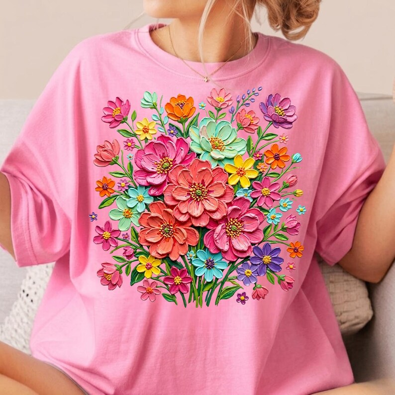 May include: A pink t-shirt featuring a vibrant floral design. The bouquet includes large, textured flowers in shades of pink, coral, orange, yellow, blue, and purple, with green leaves. The shirt has short sleeves and a crew neck.