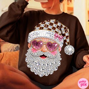 May include: A dark brown long-sleeve sweatshirt featuring a graphic of a Santa Claus face covered in sparkling rhinestones. The Santa wears a chequered hat and pink heart-shaped glasses. The design includes a small pink bow on the hat.