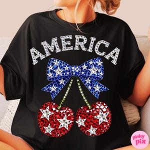 May include: Black t-shirt with the word "AMERICA" in white, sparkling letters. A blue bow with white stars and two red cherries with white stars complete the design. The cherries have green stems. A patriotic themed graphic tee.