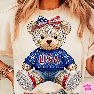 May include: A cream-colored t-shirt featuring a bedazzled teddy bear design. The bear wears a blue sweater with "USA" in red, denim shorts, and shoes with red and white accents. A bow with a star and stripe pattern sits atop the bear's head.