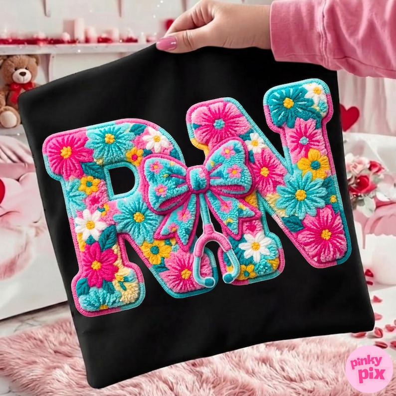 May include: Black t-shirt featuring the letters "RN" in a floral design. The letters are filled with pink, blue, yellow, and white flowers. A bow and stethoscope are in the center.