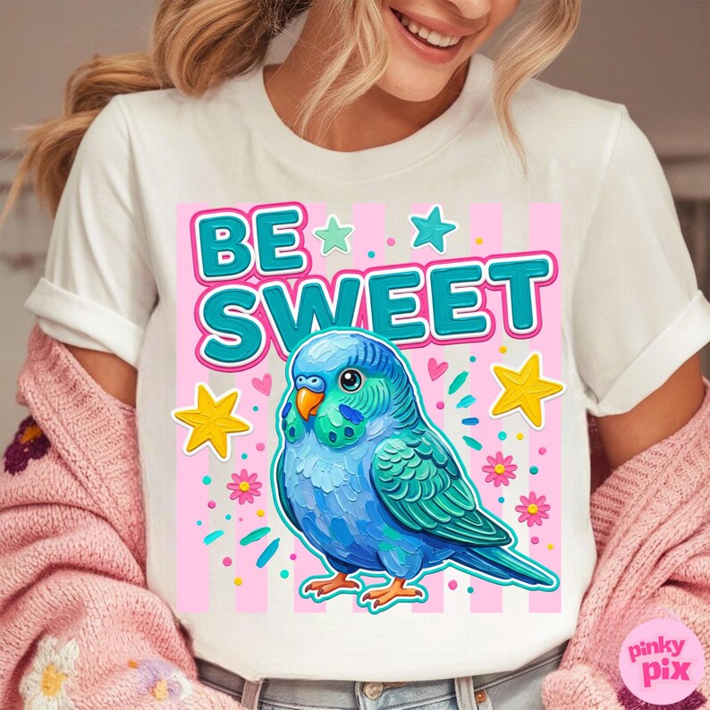 May include: White t-shirt featuring a colorful illustration of a blue parakeet with the text "BE SWEET" in teal and pink. The design includes stars and flowers, creating a cheerful and playful aesthetic.