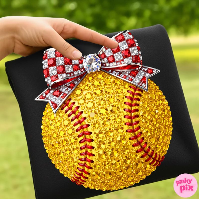 May include: A bedazzled softball with a red and white bow on a black surface. The softball is covered in yellow gems with red stitching. The bow features red and white checkered accents and a large, clear gem in the centre.